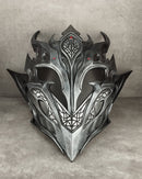 A detailed fantasy metal helmet with sharp angular spikes, intricate geometric engravings, and small red gem accents, displayed against a textured gray concrete background.