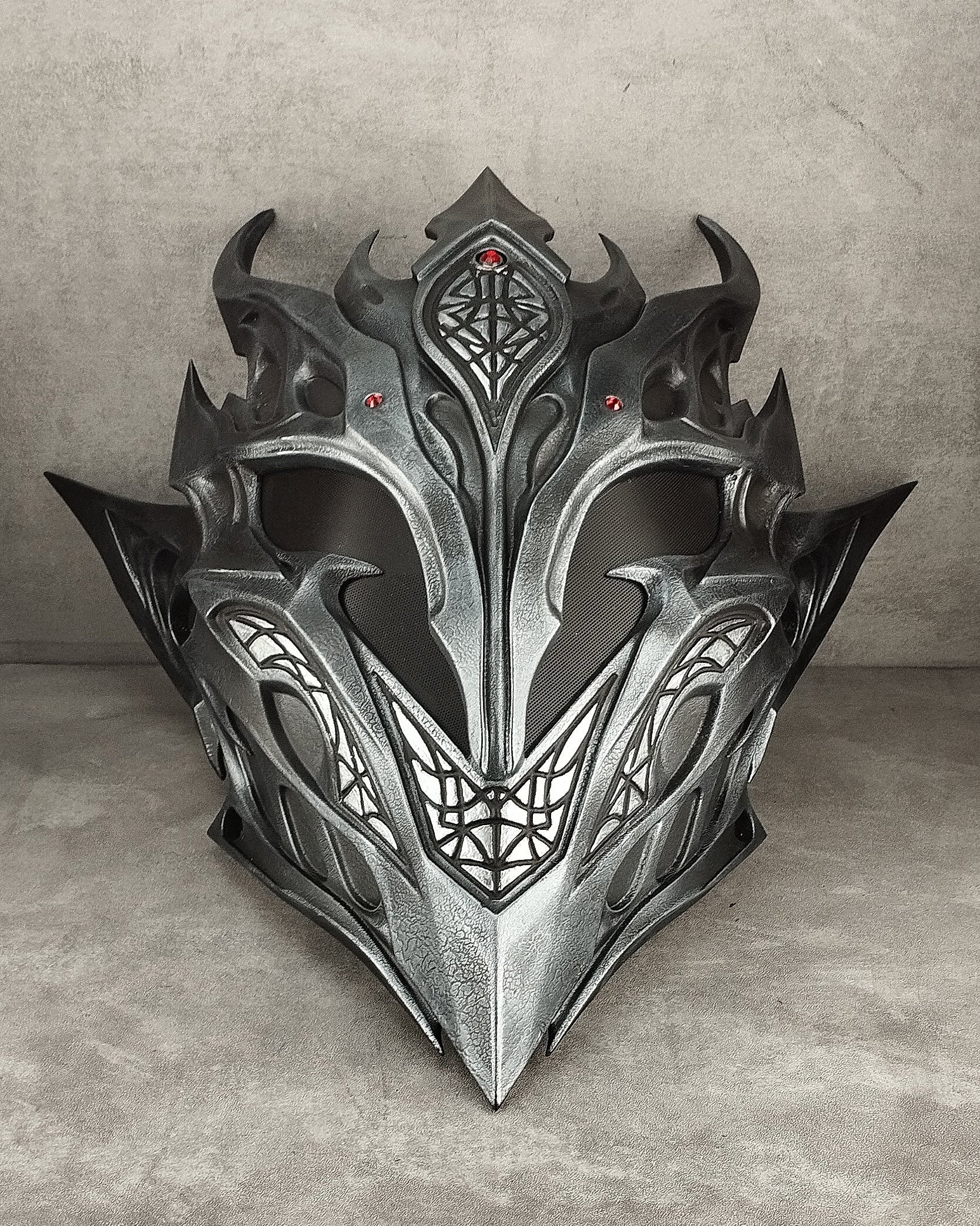 A detailed fantasy metal helmet with sharp angular spikes, intricate geometric engravings, and small red gem accents, displayed against a textured gray concrete background.