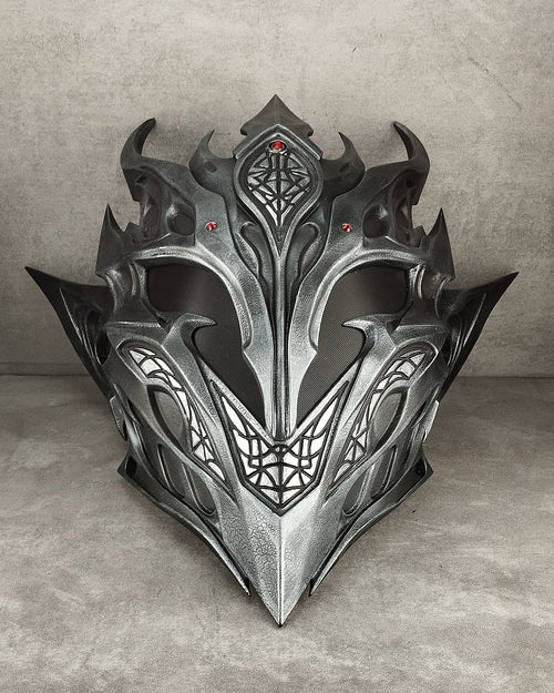 A detailed fantasy metal helmet with sharp angular spikes, intricate geometric engravings, and small red gem accents, displayed against a textured gray concrete background.
