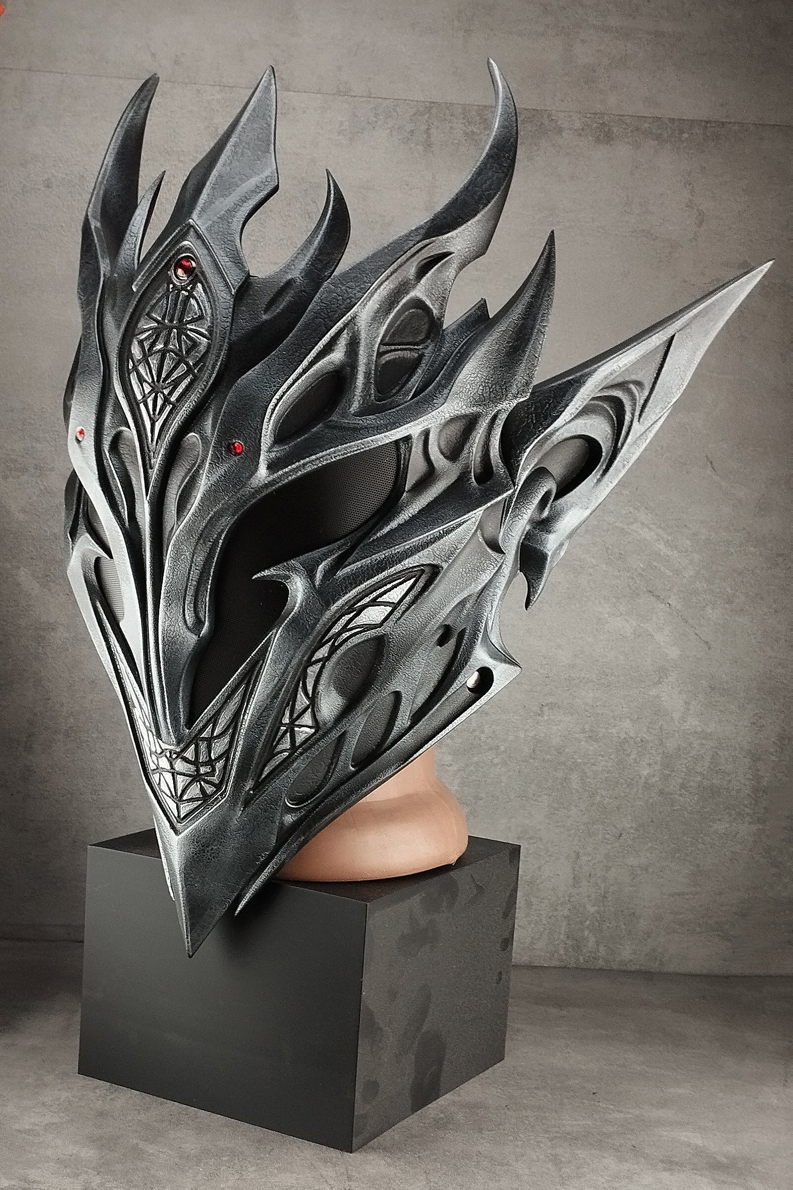 A highly detailed, metallic silver and black fantasy helmet with sharp flame-like spikes and intricate carvings, displayed on a black pedestal against a textured gray studio background.