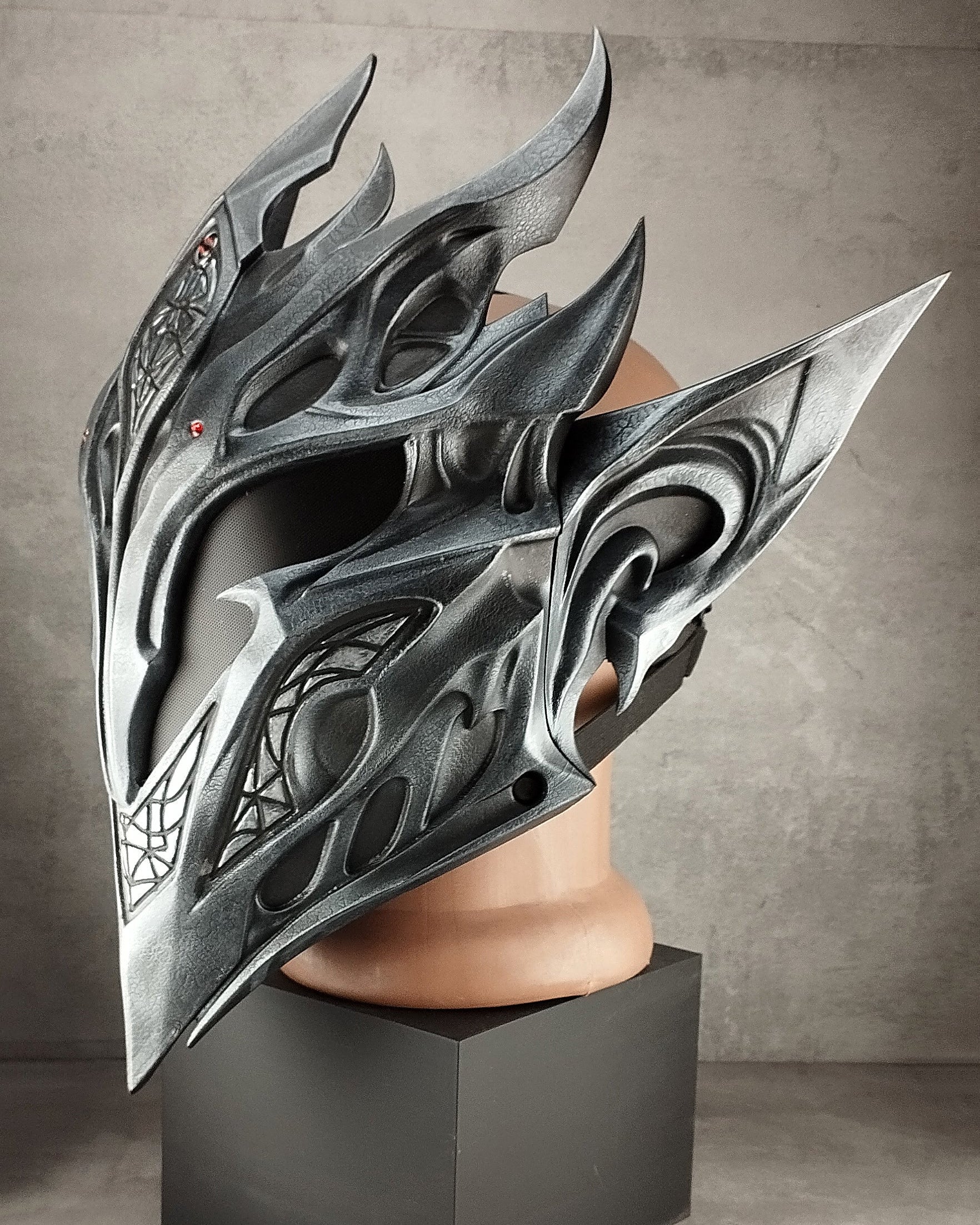 A large, ornate metallic fantasy helmet displayed on a mannequin head mounted in a black pedestal against a textured gray studio background.