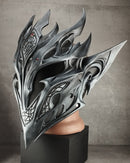 A highly detailed, metallic silver fantasy helmet with sharp, flame-like spikes and intricate dark engravings, displayed on a black pedestal stand against a textured gray concrete wall.