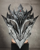 A large, ornate fantasy-style metallic mask with sharp angular horns and intricate geometric detailing, displayed on a black pedestal against a textured gray wall.