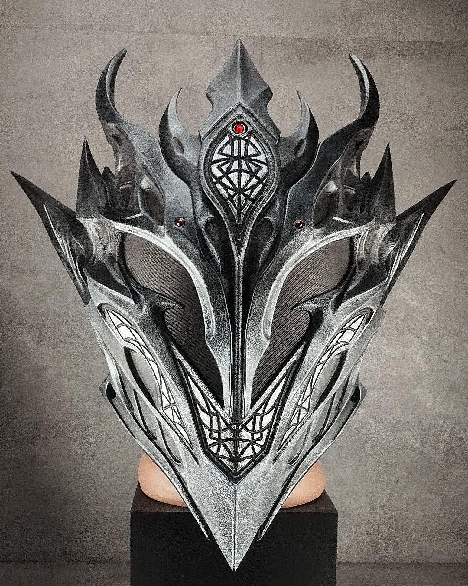 A large, ornate fantasy-style metallic mask with sharp angular horns and intricate geometric detailing, displayed on a black pedestal against a textured gray wall.