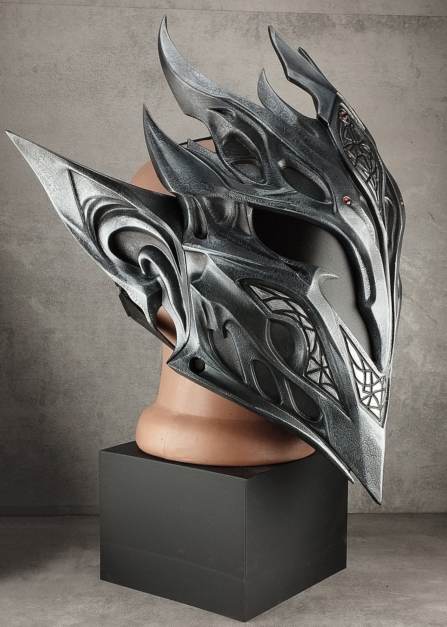 A large, ornate fantasy-style helmet with sharp angular spikes and dark metallic detailing, displayed on a mannequin head mounted atop a black pedestal against a textured gray wall.
