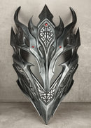 A metallic silver and black fantasy dragon mask with sharp horns and intricate geometric engravings, displayed against a textured gray concrete wall and floor.
