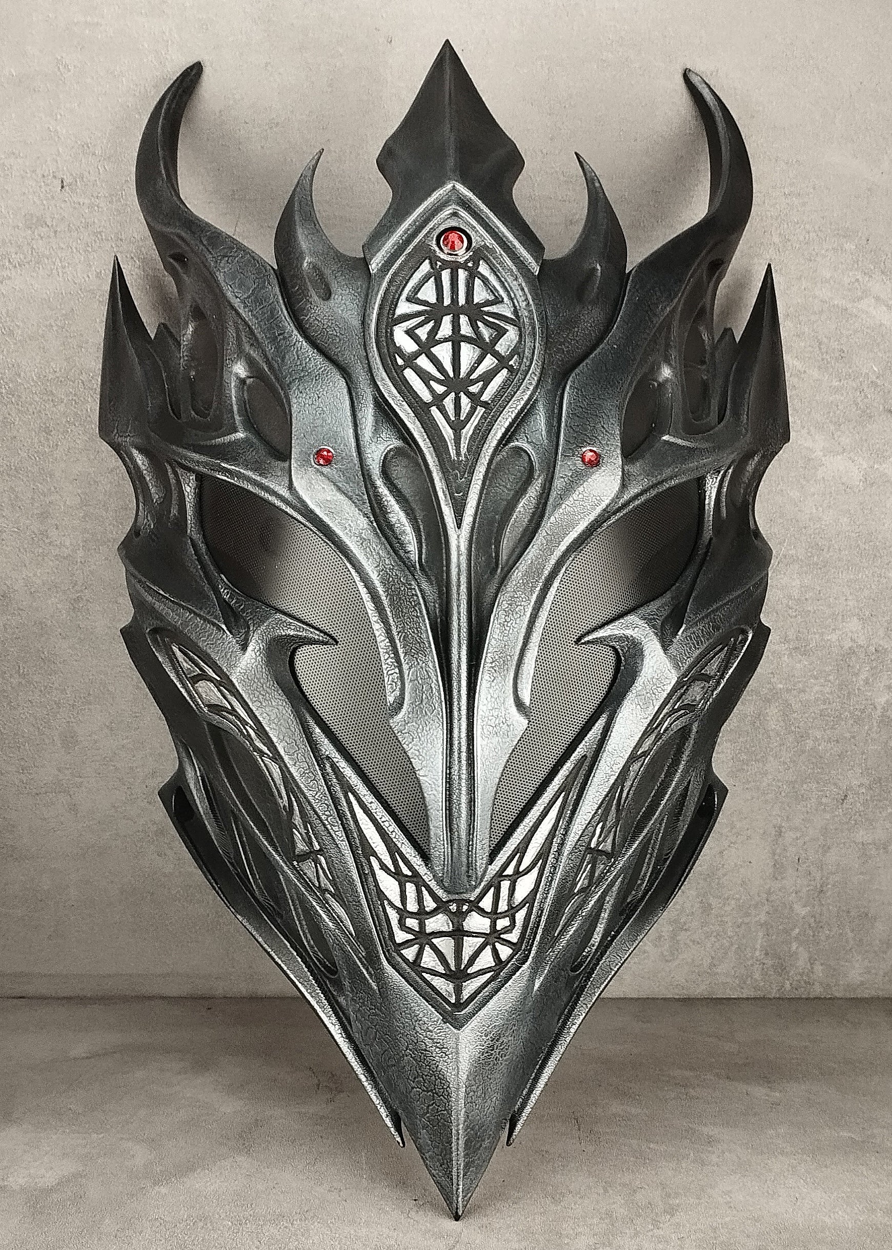 A metallic silver and black fantasy dragon mask with sharp horns and intricate geometric engravings, displayed against a textured gray concrete wall and floor.