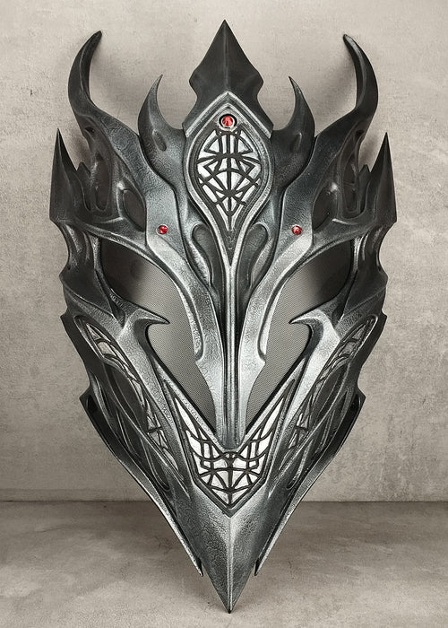 A metallic silver and black fantasy dragon mask with sharp horns and intricate geometric engravings, displayed against a textured gray concrete wall and floor.