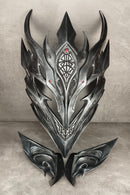 A dark metallic fantasy mask with sharp angular horns and symmetrical wing-like side pieces, displayed against a textured gray backdrop.