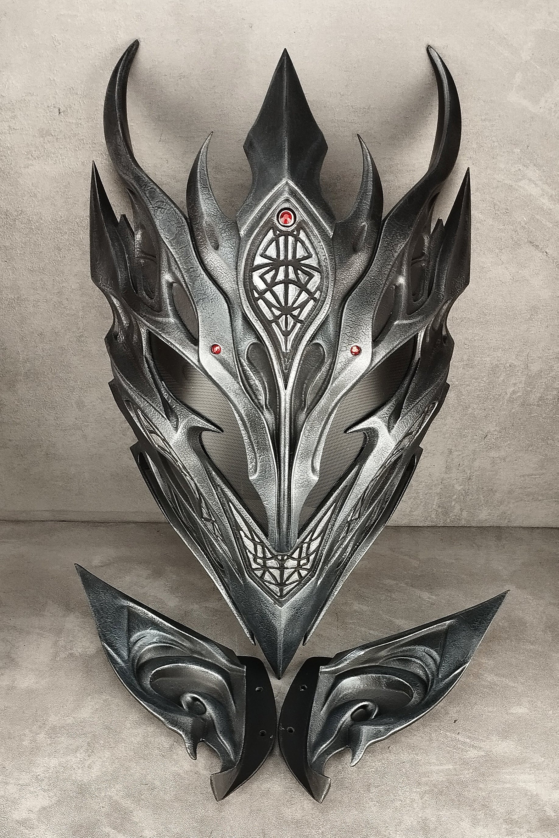 A dark metallic fantasy mask with sharp angular horns and symmetrical wing-like side pieces, displayed against a textured gray backdrop.