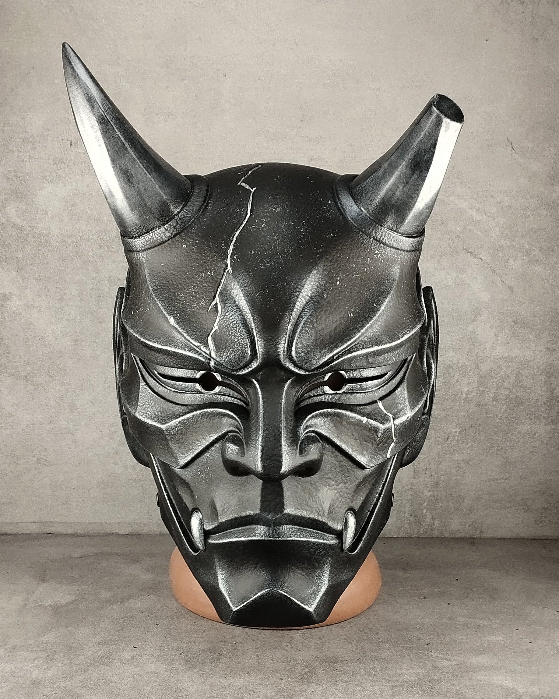 A black and silver horned demon mask displayed on a mannequin head against a gray concrete wall and floor.