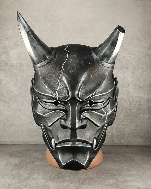 A black and silver horned demon mask displayed on a mannequin head against a gray concrete wall and floor.