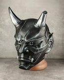 A black and silver horned demon mask displayed on a brown mannequin head against a gray concrete wall.