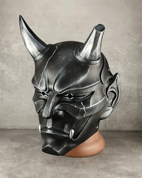 A black and silver horned demon mask displayed on a brown mannequin head against a gray concrete wall.