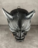 A dark metallic gothic horned mask with a cracked forehead design, displayed against a textured gray concrete wall and floor.