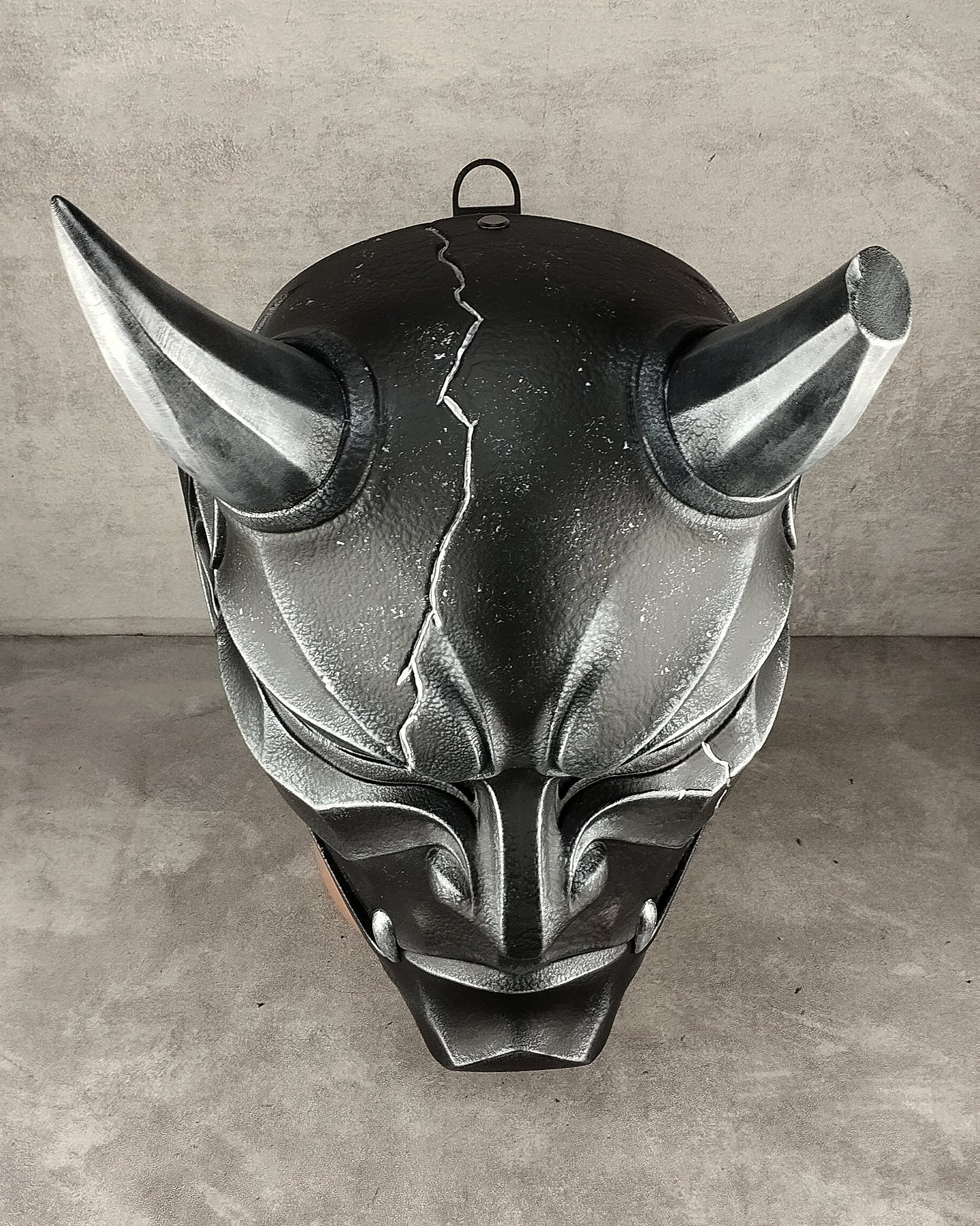 A dark metallic gothic horned mask with a cracked forehead design, displayed against a textured gray concrete wall and floor.