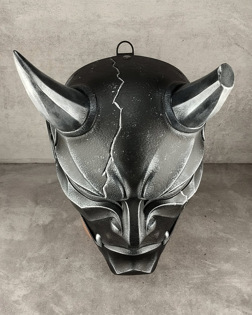 A dark metallic gothic horned mask with a cracked forehead design, displayed against a textured gray concrete wall and floor.
