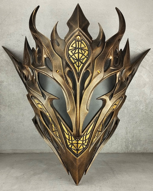 A highly detailed fantasy elf mask with sharp horns, metallic gold and bronze scales, and intricate geometric engravings, set against a textured gray concrete background.