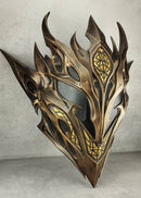 A metallic bronze and gold fantasy mask with sharp flame-like spikes and intricate geometric engravings, displayed against a textured gray concrete wall.
