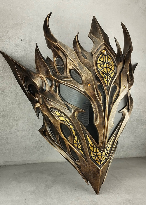 A metallic bronze and gold fantasy mask with sharp flame-like spikes and intricate geometric engravings, displayed against a textured gray concrete wall.