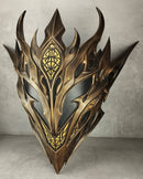 A metallic bronze and gold fantasy elf mask with sharp, flame-like horns and intricate geometric engravings, displayed against a textured gray concrete wall and floor.