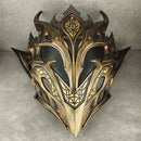 A fantasy-style metallic elf mask with ornate gold and black detailing, photographed against a gray concrete wall and floor.