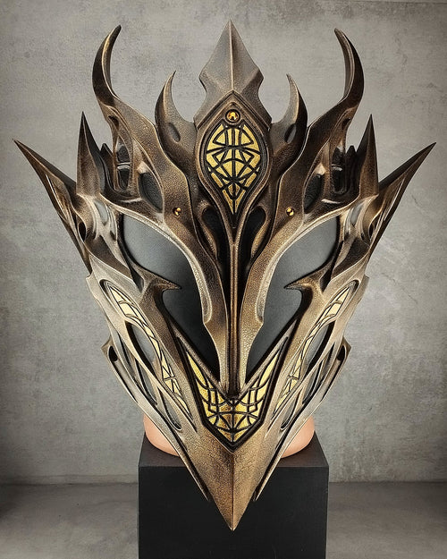 A large ornate fantasy-style elf mask with sharp metallic spikes and gold detailing, displayed on a black pedestal in a gray indoor setting.