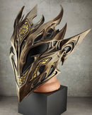 A highly detailed fantasy elf mask with sharp metallic gold and bronze wings, displayed on a black pedestal against a gray concrete wall.