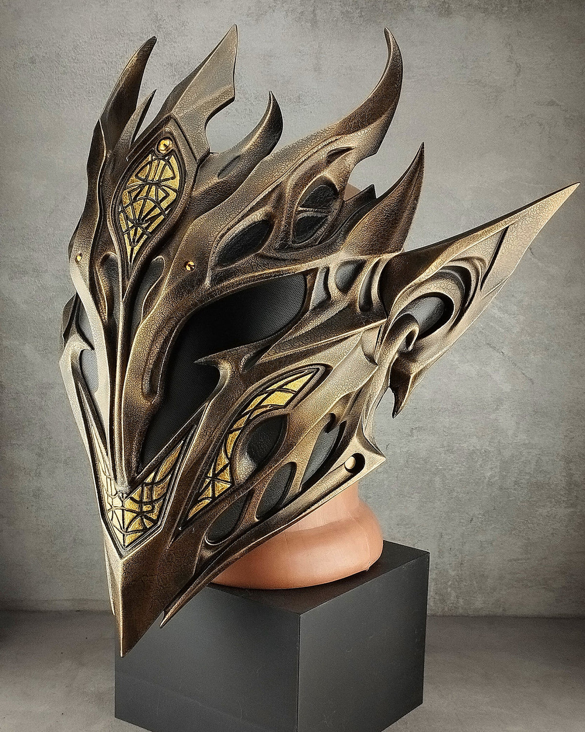 A highly detailed fantasy elf mask with sharp metallic gold and bronze wings, displayed on a black pedestal against a gray concrete wall.