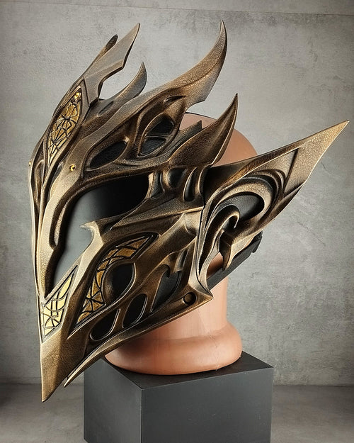 A large ornate fantasy-style elf mask with metallic bronze and black flame-like detailing, displayed on a tan mannequin head mounted on a black pedestal against a gray studio backdrop.
