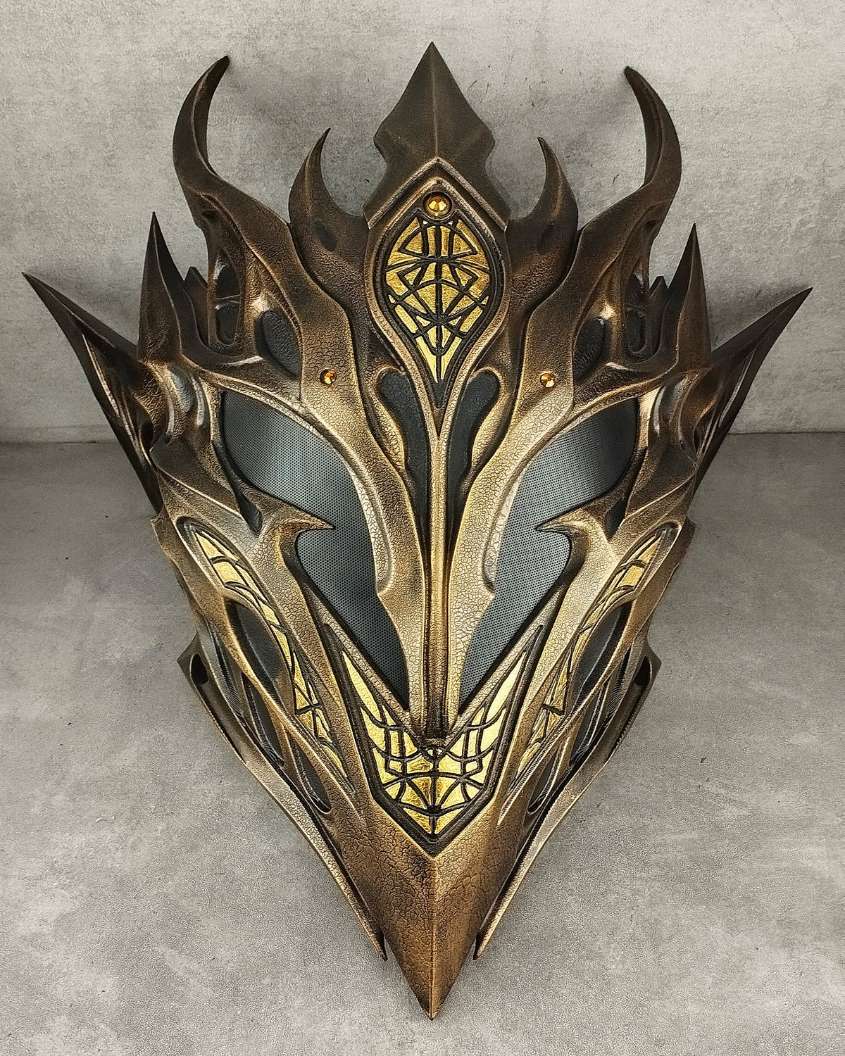 A fantasy-style metallic elf mask with ornate pointed spikes, gold-toned detailing, and dark recessed areas, photographed against a textured gray wall and floor.