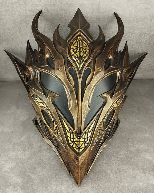 A fantasy-style metallic elf mask with ornate pointed spikes, gold-toned detailing, and dark recessed areas, photographed against a textured gray wall and floor.