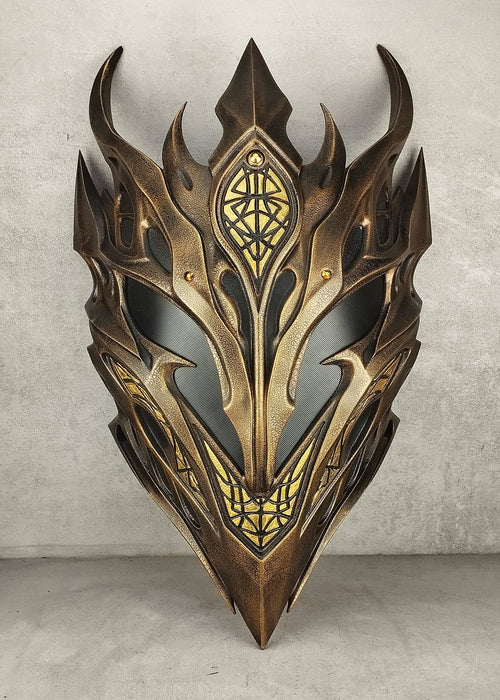 A fantasy-style metallic elf mask with sharp angular horns and ornate gold detailing, photographed against a textured light gray wall and floor.