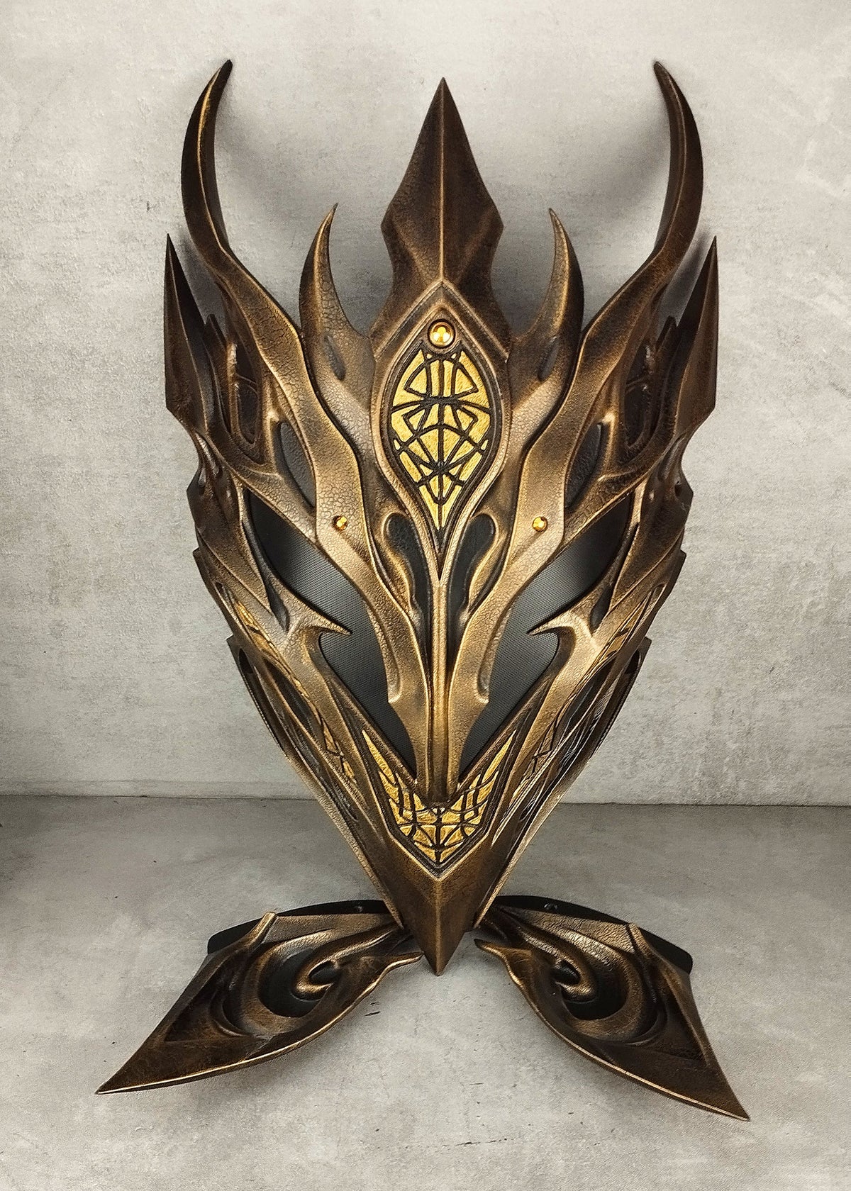 A fantasy-style metallic elf mask  with sharp angular horns and ornate gold detailing, photographed standing against a textured gray wall and floor.