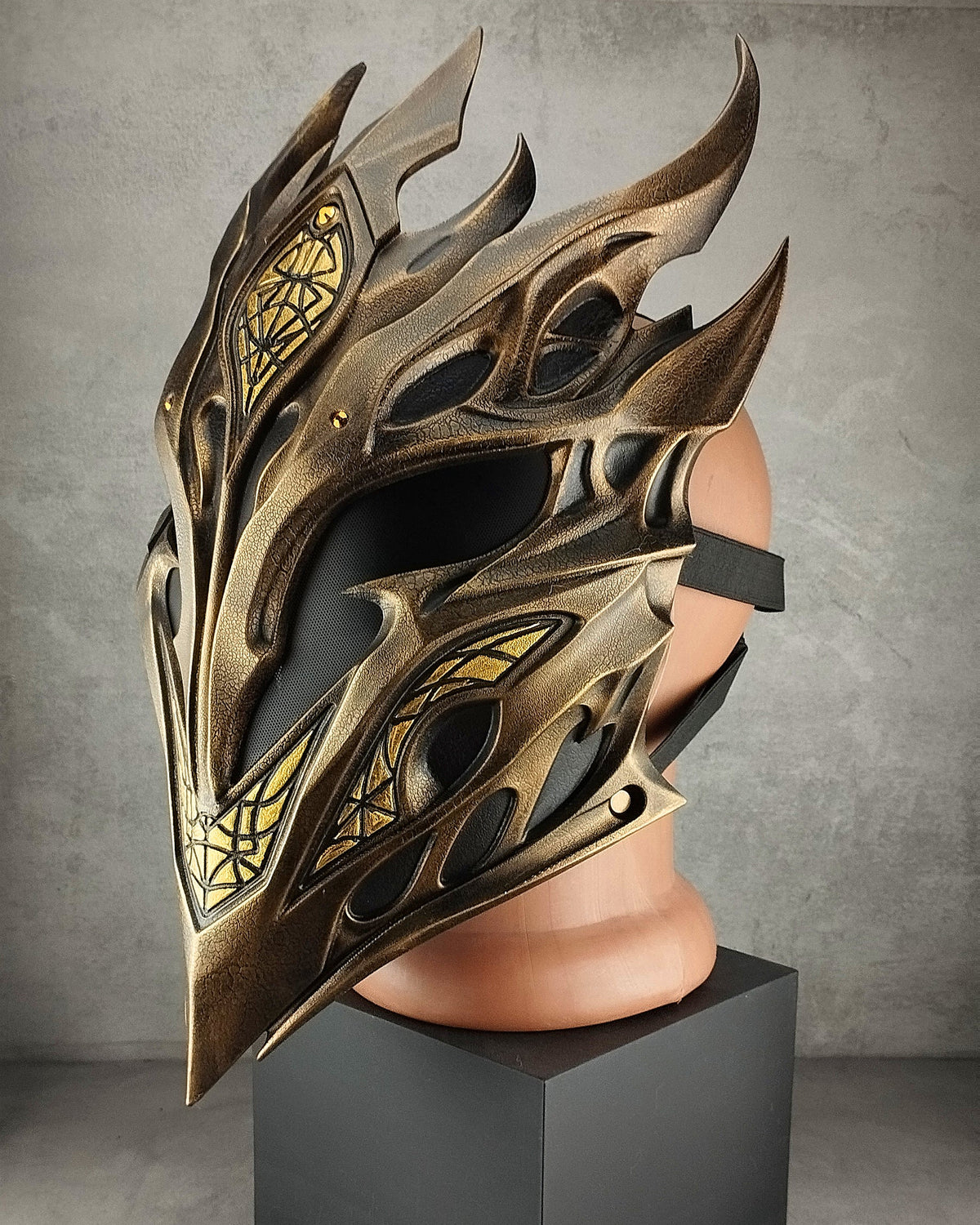 A metallic fantasy-style elf mask with sharp angular spikes and gold decorative linework, displayed on a flesh-toned mannequin head mounted over a black pedestal in a gray studio setting.