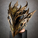 A highly detailed fantasy mask with sharp, flame-like metallic spikes and intricate cutouts, finished in a bronze-gold color with black recessed areas and small gold accents. The mask is displayed on a black pedestal against a textured gray studio background.